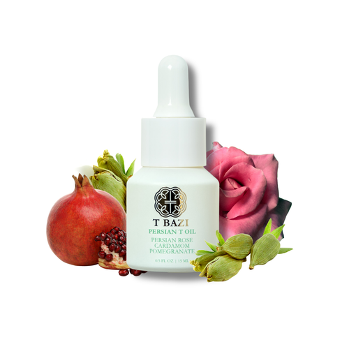A white dropper bottle of T Bazi Persian Rose Oil, styled with fresh ingredients including a pomegranate, rose petals, and cardamom pods on a solid white background.