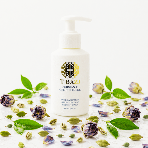 A bottle of T Bazi Persian T Gel Cleanser standing upright on a white surface, surrounded by green tea leaves, dried flower buds, and scattered petals against a clean white background.