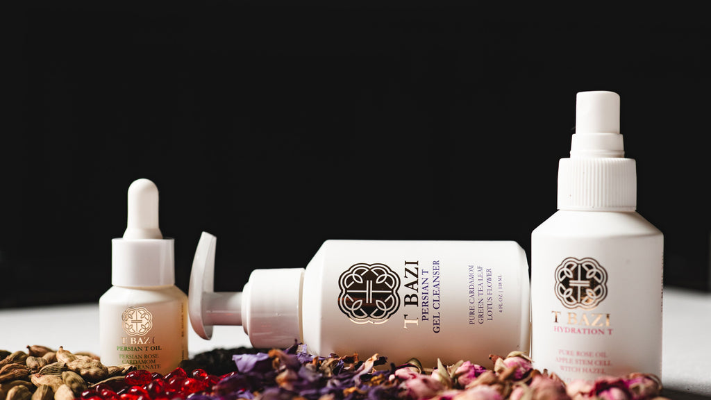 Three white skincare bottles from the brand T. Bazi, including a pump bottle, a dropper bottle, and a spray bottle, arranged on a white surface with dried flowers and botanicals against a black background.